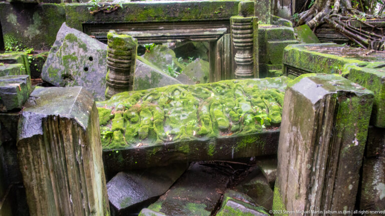 Moss-covered carved lintel lying among fallen sandstone blocks, pillars, and surviving openings.