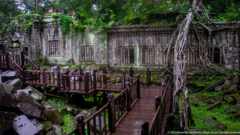 Raised wooden walkways crossing rubble before a rooted sandstone gallery with baluster windows.