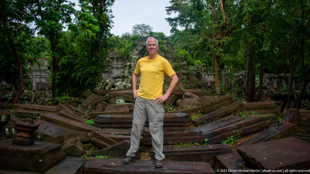 Man in a yellow shirt standing among collapsed temple blocks with a ruined gateway behind him.