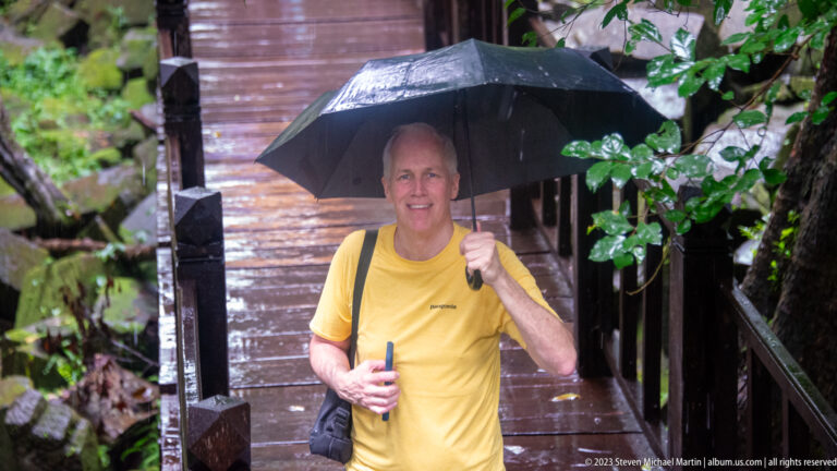 Man in yellow holding a black umbrella on a wet wooden walkway among temple ruins.