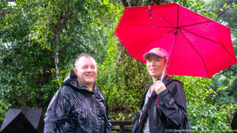 Two visitors in dark rain jackets standing beneath a bright red umbrella in lush forest.