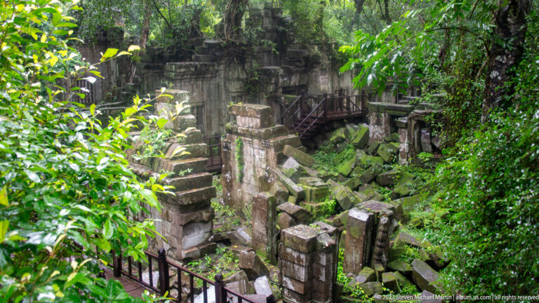 Elevated view of a ruined gateway with timber stairs, walkways, foliage, and fallen sandstone.