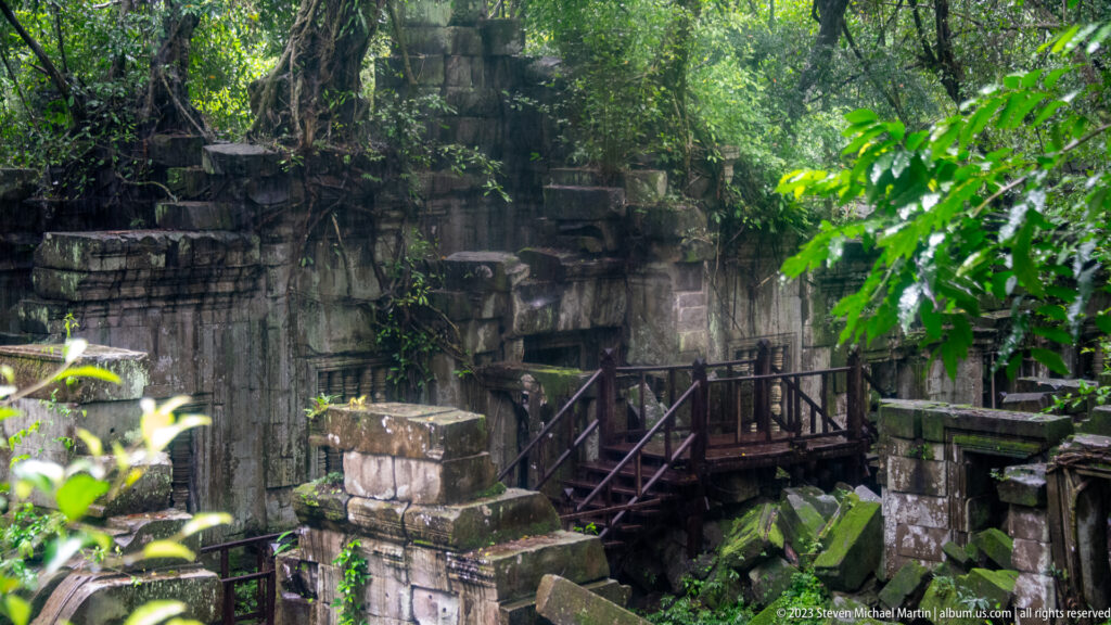 Wooden bridge crossing between dark sandstone ruins, collapsed blocks, and dense green vegetation.