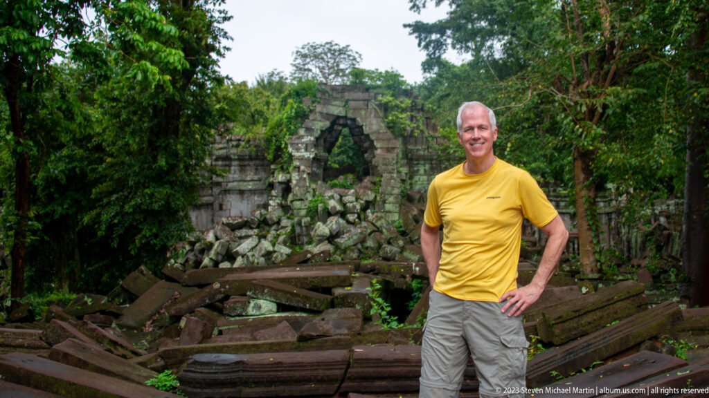 Man in yellow posing before a pointed ruined doorway and scattered sandstone beams.