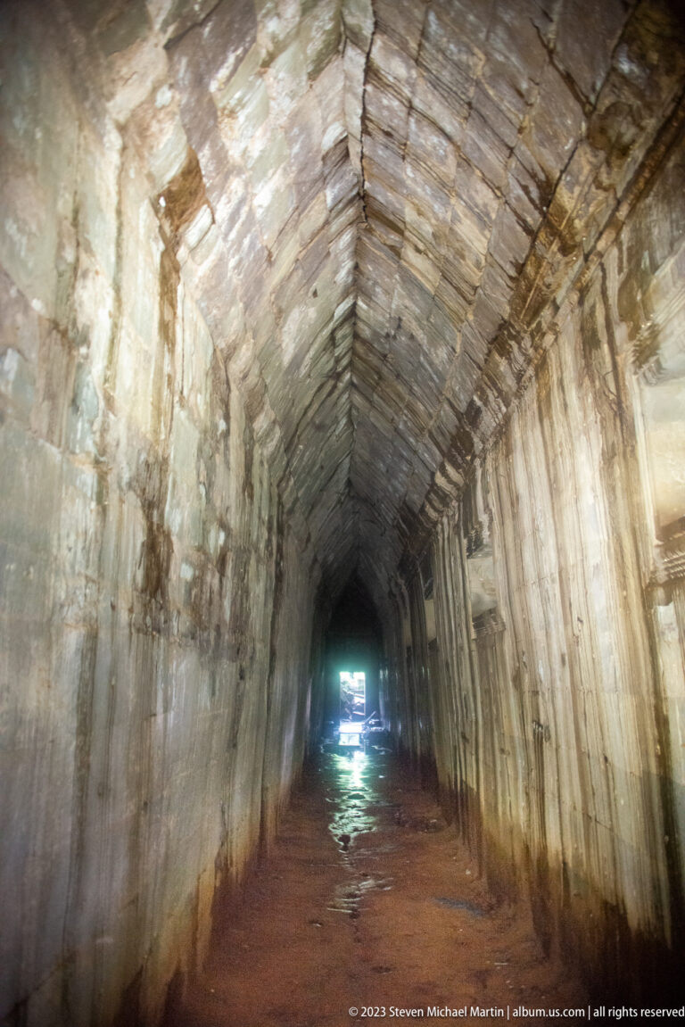 Long sandstone corridor with a pointed corbel vault, stained walls, and a wet floor.