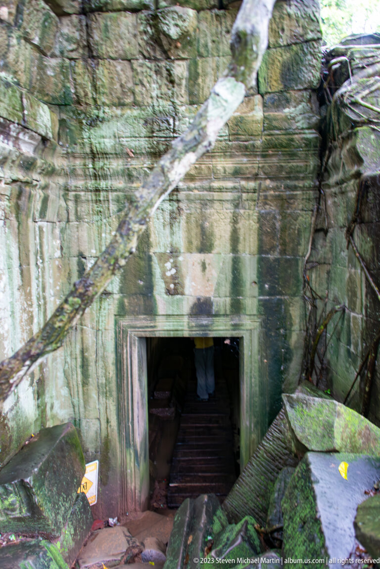 Low sandstone doorway leading into a dark passage with roots, rubble, and a wooden walkway.