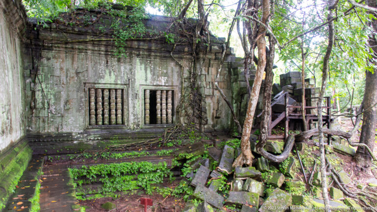 Sandstone gallery with two baluster windows, mossy base, trees, roots, and collapsed masonry.