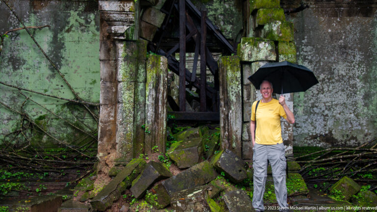 Man with black umbrella standing beside a timber-braced doorway amid fallen sandstone and trees.