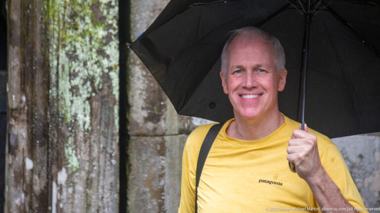 Smiling man in yellow beneath a black umbrella against a weathered sandstone wall.