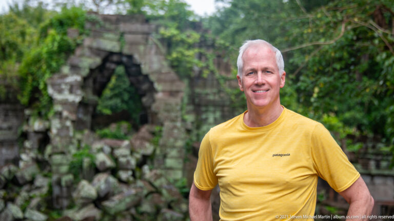 Waist-up portrait of a smiling man in yellow with a blurred ruined gateway behind him.