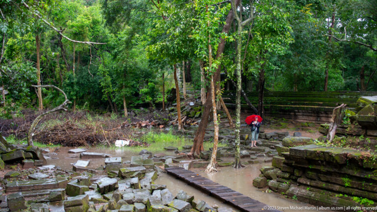 Wide flooded ruin field with trees, sandstone blocks, timber path, and a distant red umbrella.