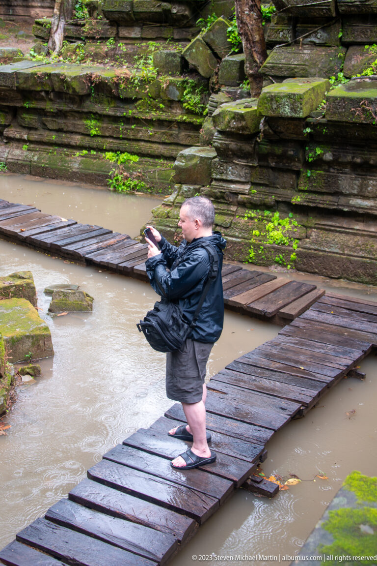 Visitor photographing from wet timber boards above shallow floodwater beside sandstone walls.