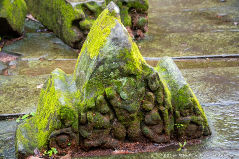 Moss-covered triangular sandstone architectural fragment with worn carved ornament resting on wet temple paving.