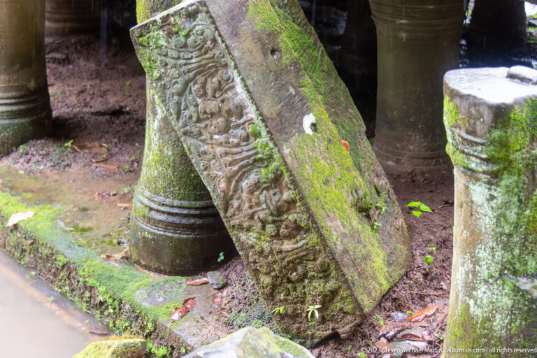 Mossy carved sandstone lintel leaning diagonally against large turned columns inside the ruined temple.
