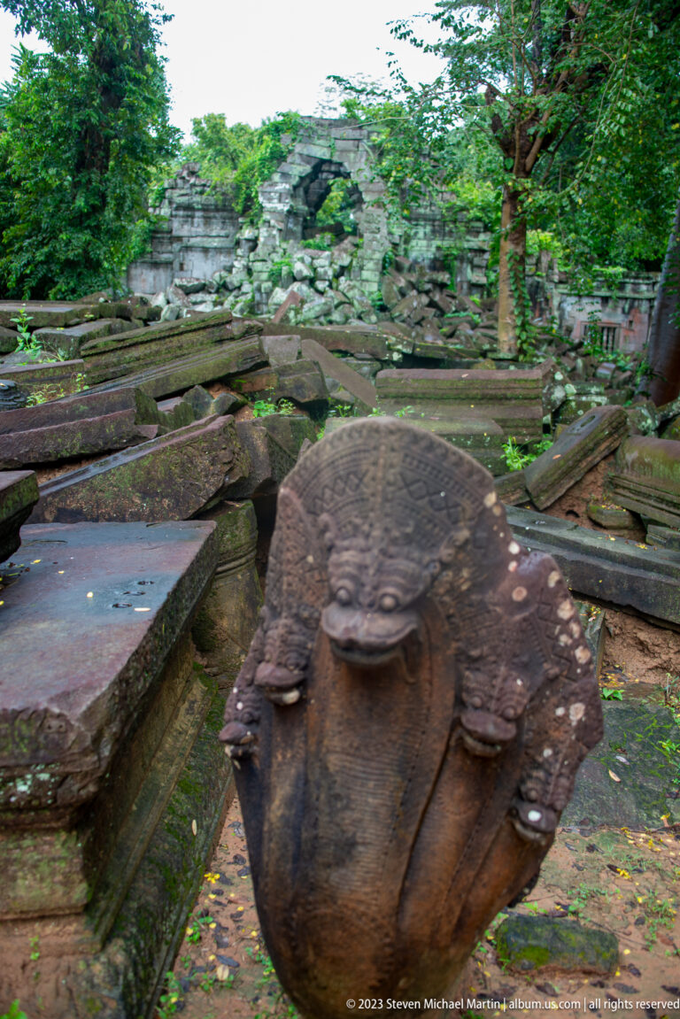 Weathered multi-headed naga sculpture in front of long fallen sandstone blocks and temple ruins.