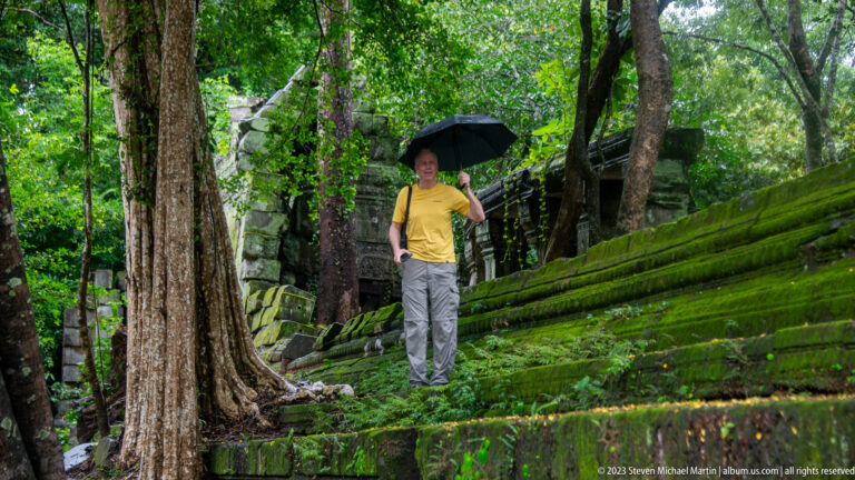 Man with black umbrella descending steep moss-covered sandstone steps among trees and ruins.
