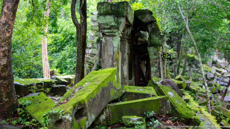 Partly standing sandstone doorway surrounded by large moss-covered fallen blocks in forest.