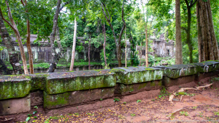 Low moss-covered sandstone wall extending through forest with temple galleries visible beyond.