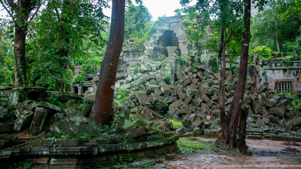 Forest view of a surviving stone gateway above heaps of mossy collapsed masonry.