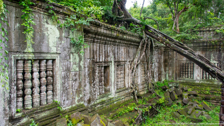 Oblique view of a mossy sandstone gallery with repeated balustered windows, fallen blocks, and a leaning tree.