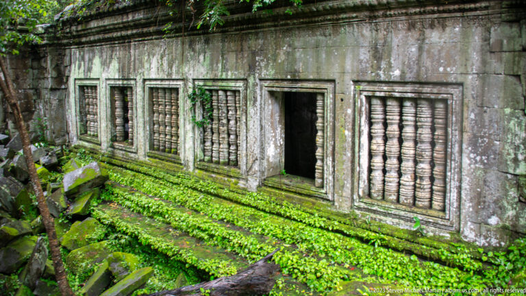 Front view of a sandstone gallery with four balustered windows, an open doorway, moss, and fallen stones.
