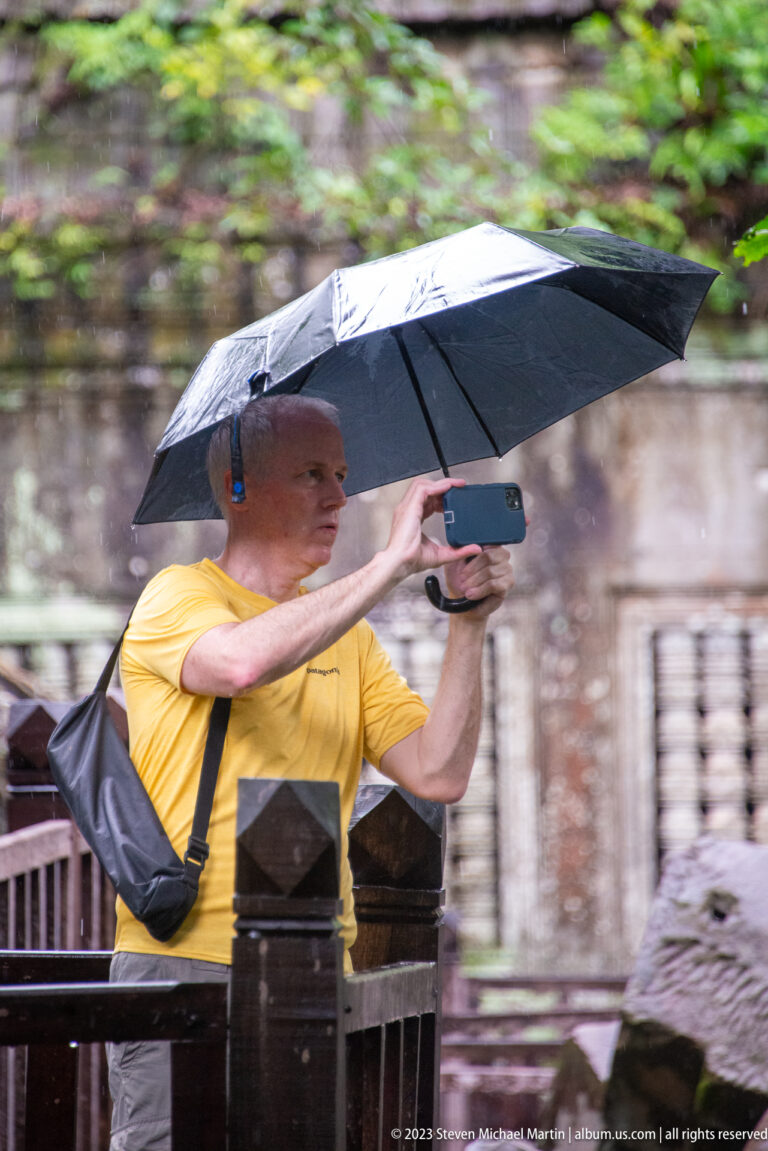 Visitor in a yellow shirt holds an umbrella and smartphone on a wet wooden walkway at Beng Mealea.