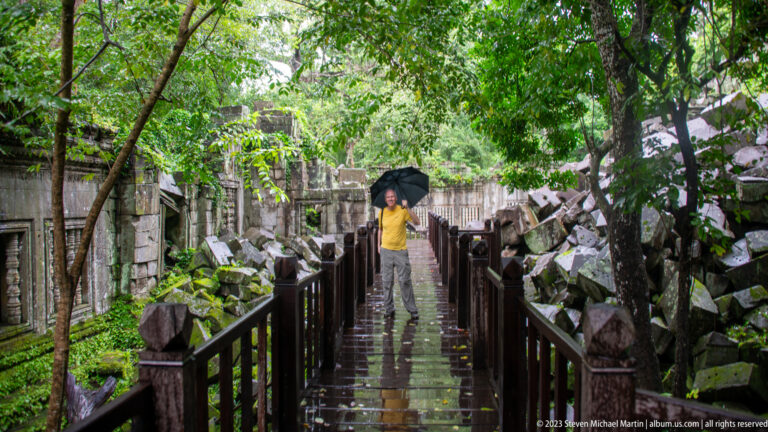 Wet wooden walkway passes between collapsed sandstone blocks and forest, with an umbrella-carrying visitor ahead.