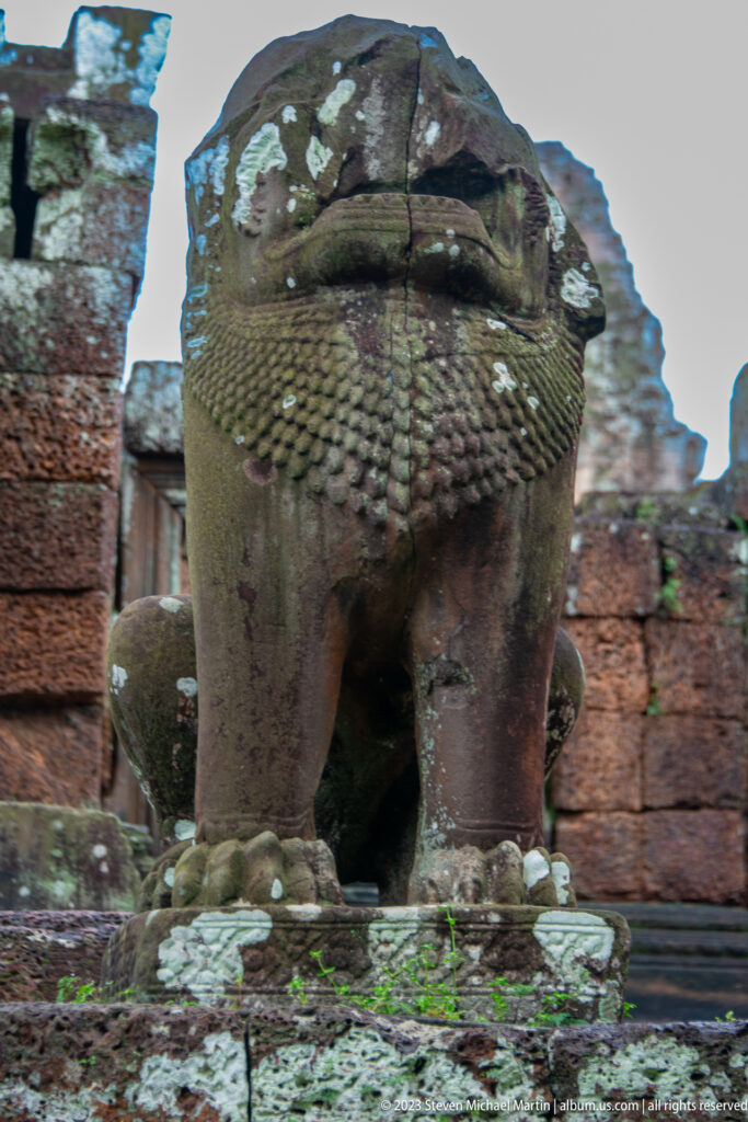 Low frontal close-up of a weathered stone lion guardian with lichen, set against East Mebon masonry.