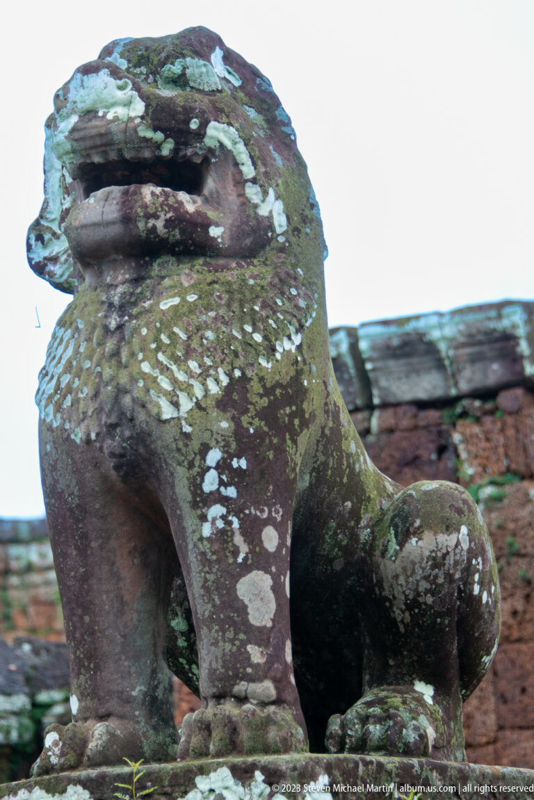 Three-quarter view of a lichen-covered stone lion guardian with raised head and curled tail at East Mebon.
