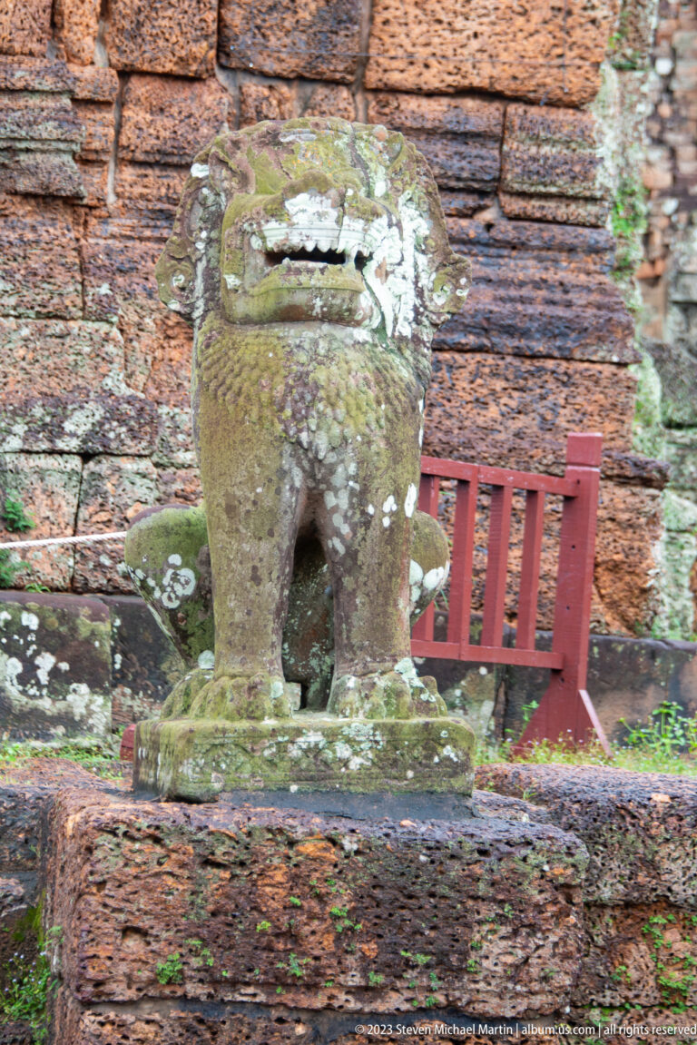 Rear view of a mossy stone lion guardian beside a red protective barrier and brick temple wall.