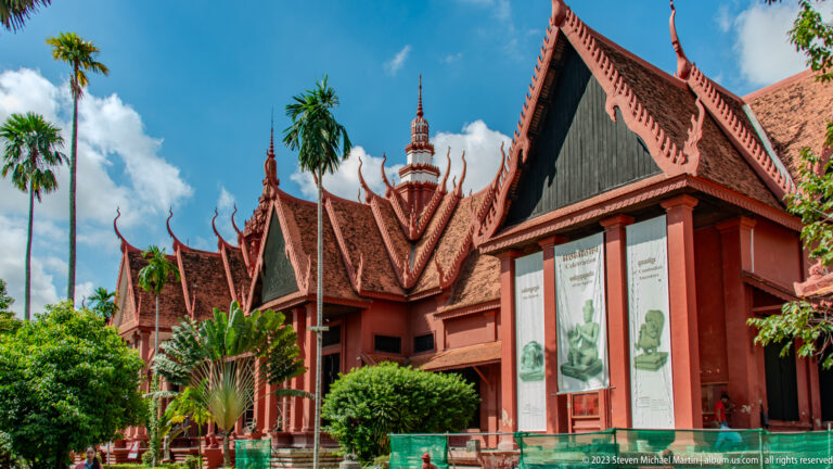 Red museum façade with tiered Khmer-style roofs, entrance banners, palms, and a central tower beneath a bright blue sky.