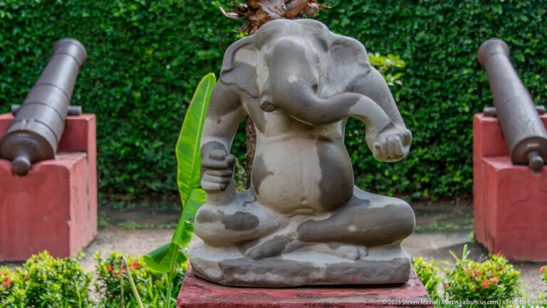 Seated stone Ganesha in a garden, framed by clipped shrubs and two dark cannon barrels.