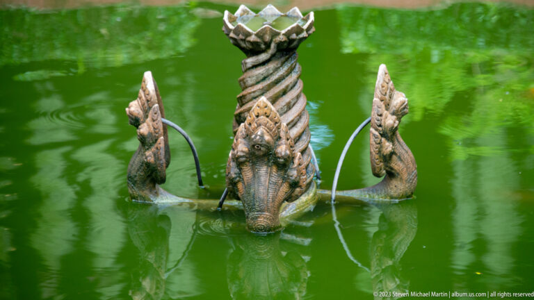 Serpent-form fountain with two water jets, a twisted central column, and a lotus-shaped bowl in a green pond.