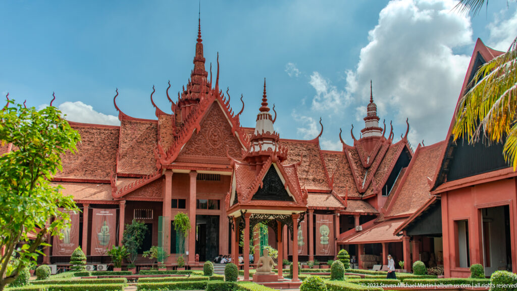 Wide courtyard view of the red museum, ornate tower, hedges, palms, and dramatic clouds.
