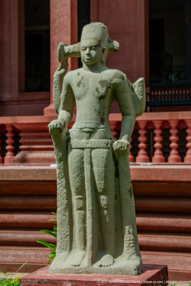 Weathered standing stone figure with bent arms positioned before the museum’s red colonnade and balustrade.