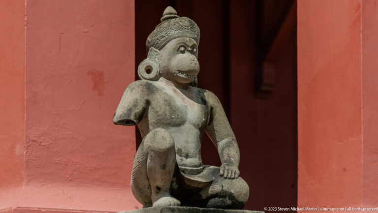 Seated monkey-faced stone figure with headdress, large ear ornaments, raised knee, and weathered surface.