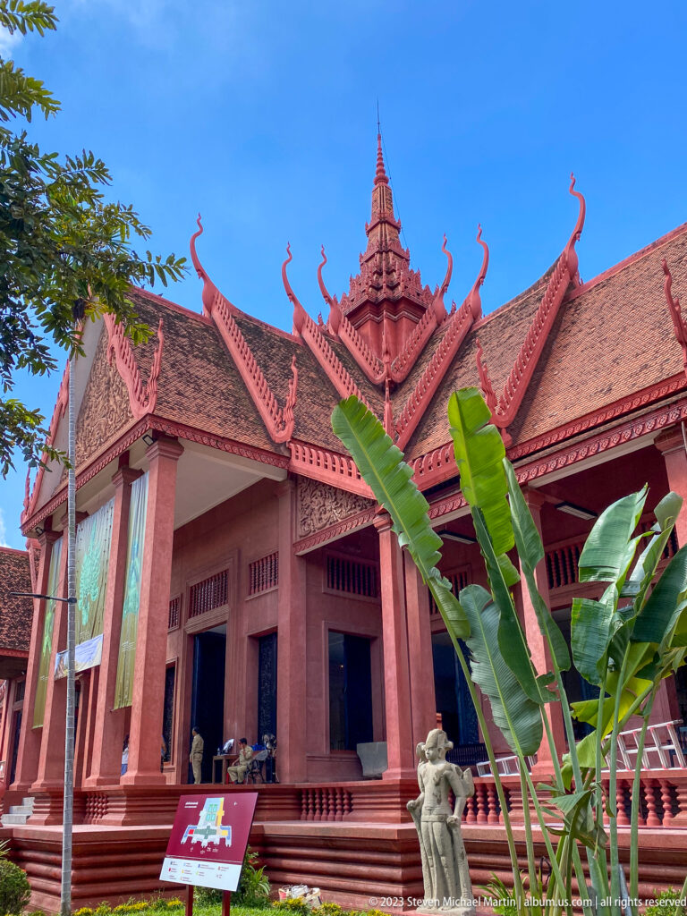 Long red museum façade with raised veranda, columns, layered roofs, banana leaves, and a small guardian figure.