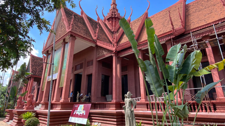 Columned red museum entrance above broad steps, framed by banana plants, a stone guardian, and blue sky.