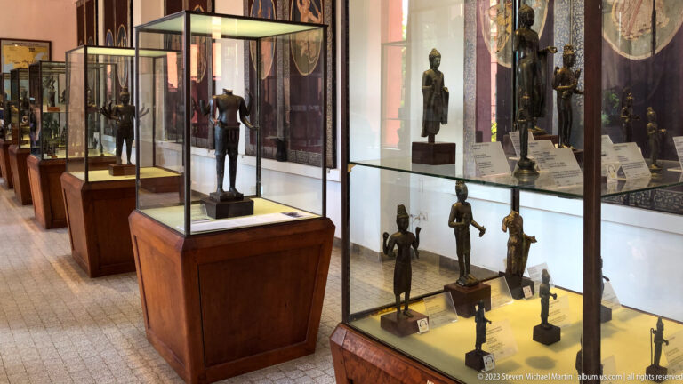 Museum gallery lined with wooden glass cases displaying standing bronze figures of varied sizes.