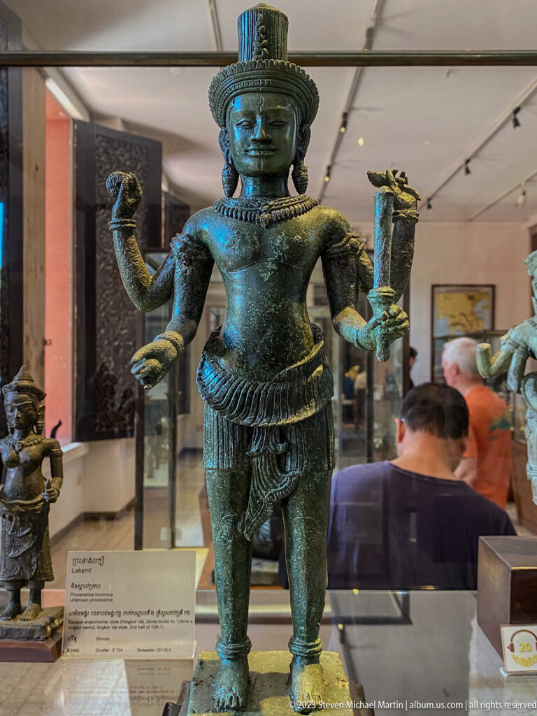 Standing four-armed bronze deity with cylindrical headdress, jewelry, patterned garment, and ritual attributes.