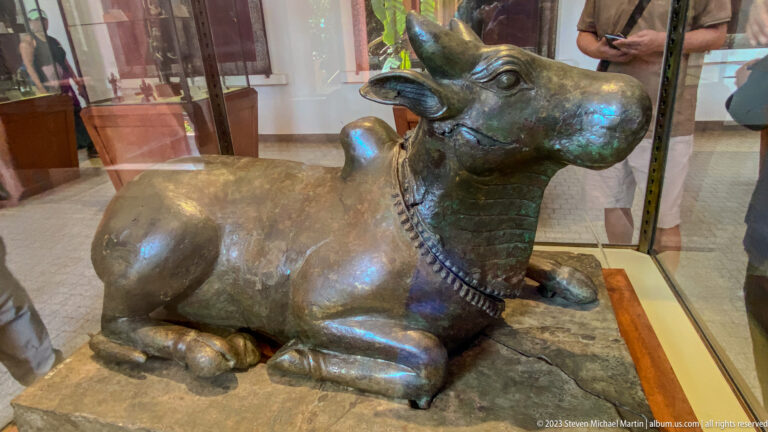 Large reclining bronze bull with curved horns, raised head, ornamental collar, and folded legs.