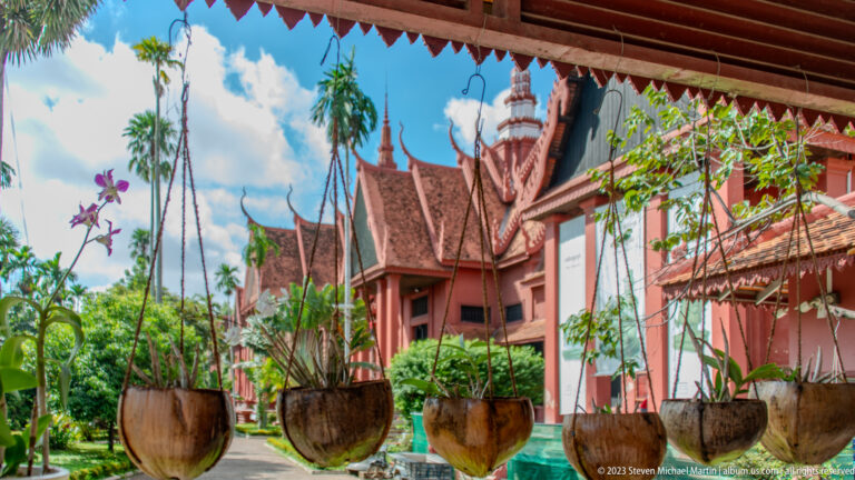 Potted tropical plants frame the red museum façade and ornate central roof tower beneath a blue sky.
