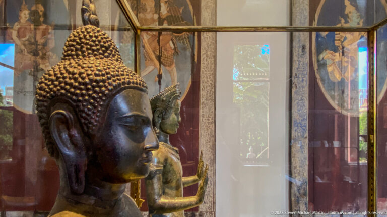 Large bronze Buddha head in profile with curled hair, rounded ushnisha, elongated earlobe, and serene expression.