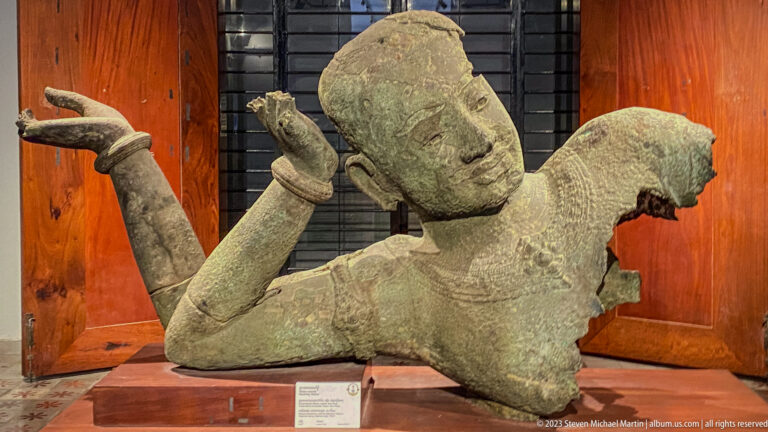 Fragmentary reclining stone figure with raised arm, jewelry, and ornate hairstyle displayed in the museum gallery.