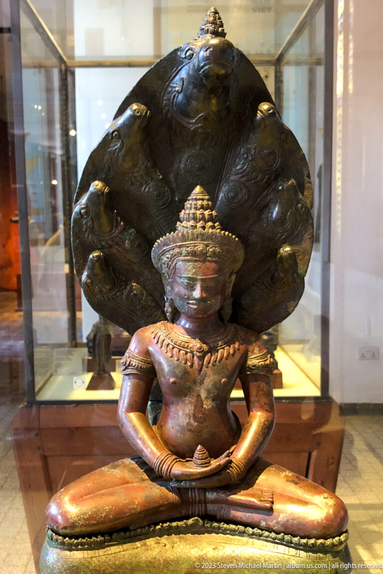 Seated bronze Buddha sheltered beneath the expanded hoods of a multi-headed naga inside a display case.