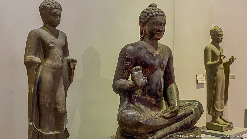 Seated Buddha with two standing Buddhist figures displayed behind it in the gallery.