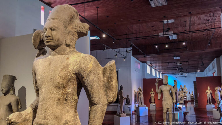 Standing sandstone figure displayed in a museum gallery with additional Khmer sculptures visible in the background.
