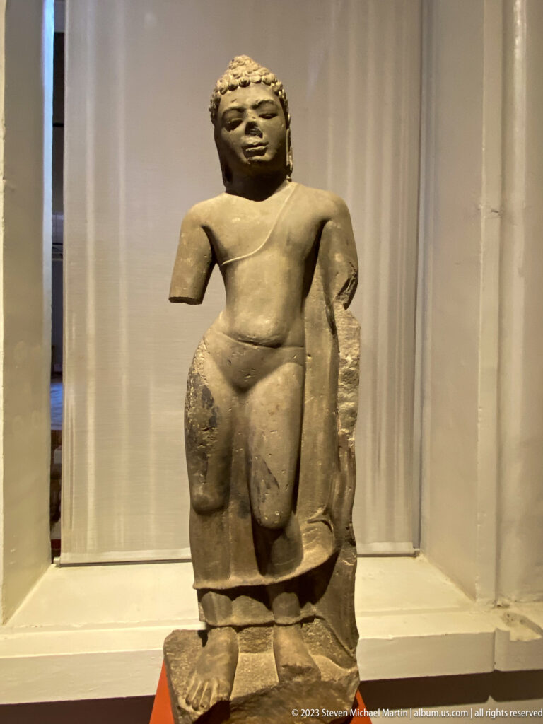 Weathered standing stone sculpture with damaged arms displayed against a light museum backdrop.