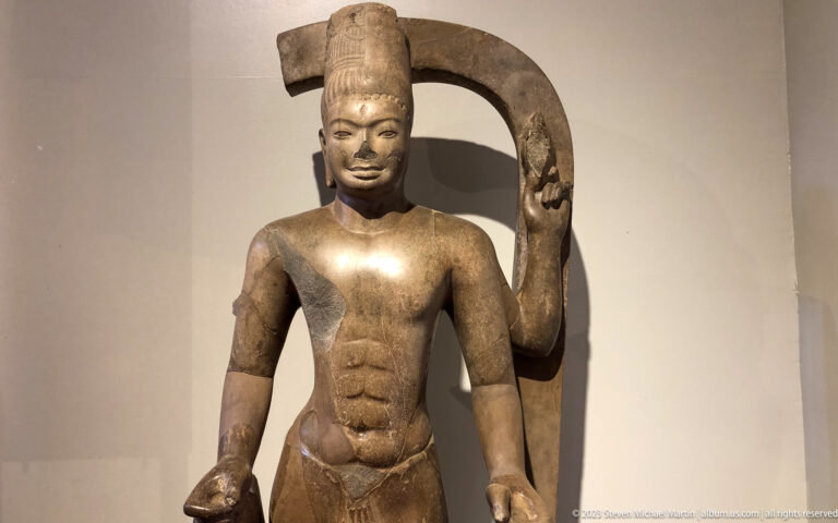 Standing crowned Khmer stone figure with elaborate jewelry and a large curved fragment behind.