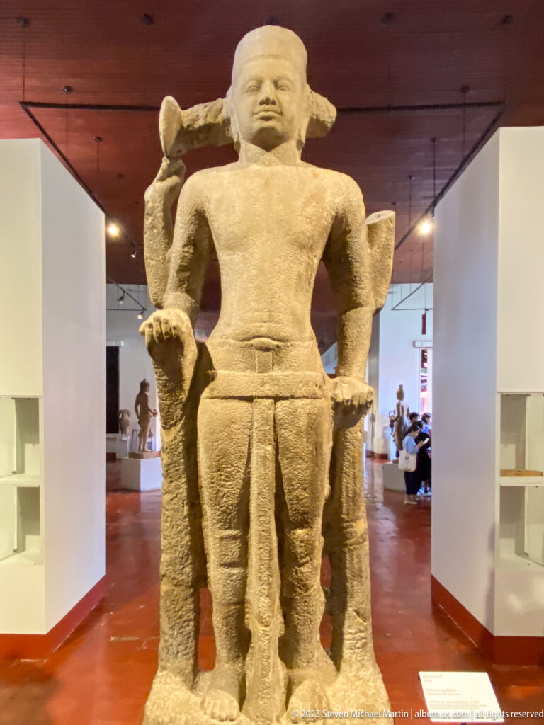 Monumental standing sandstone Khmer figure centered in a long gallery corridor with smaller sculptures beyond.